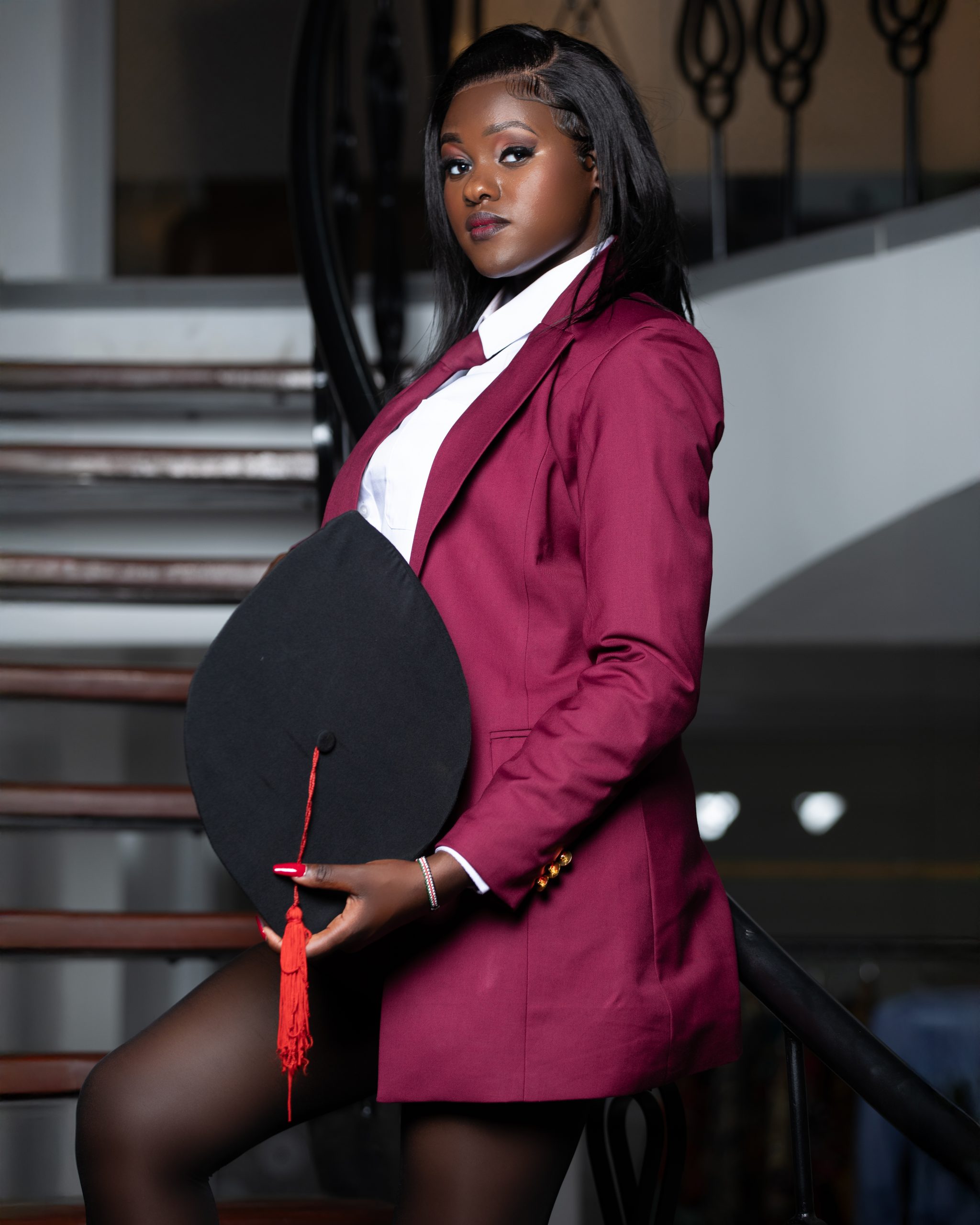 Female graduate in a maroon suit holding her graduation cap at MintGlint Studios, Nairobi.